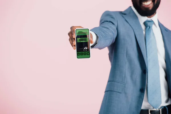 Vista recortada del hombre de negocios afroamericano en traje que muestra el teléfono inteligente con aplicación de reserva, aislado en rosa - foto de stock
