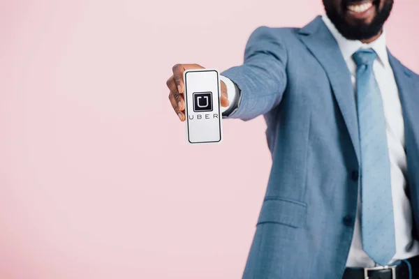 KYIV, UCRANIA - 17 de mayo de 2019: vista recortada del hombre de negocios afroamericano con traje que muestra el teléfono inteligente con la aplicación uber, aislado en rosa - foto de stock