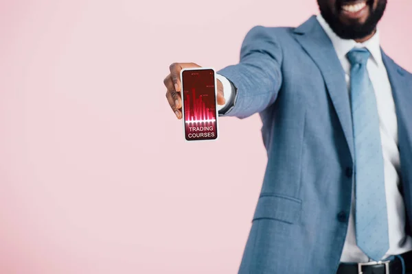 Vista recortada de hombre de negocios afroamericano en traje que muestra el teléfono inteligente con aplicación cursos de comercio, aislado en rosa - foto de stock