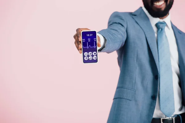 Vista recortada del hombre de negocios afroamericano en traje que muestra el teléfono inteligente con aplicación de salud, aislado en rosa — Stock Photo