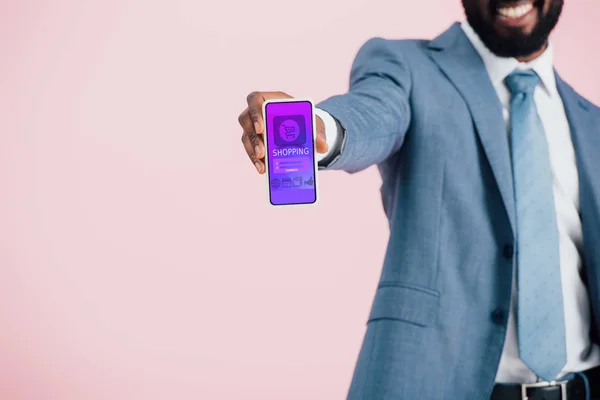 Vista recortada del hombre de negocios afroamericano en traje que muestra el teléfono inteligente con la aplicación de compras, aislado en rosa - foto de stock