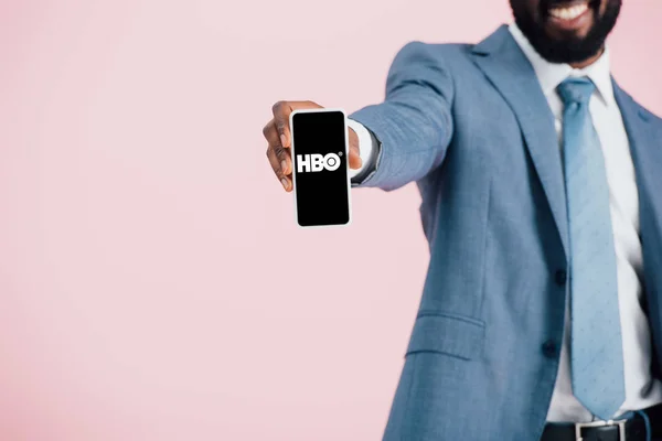 КИЕВ, УКРАИНА - 17 мая 2019 года: обрезанный вид африканского американского бизнесмена в костюме, показывающий смартфон с приложением HBO, изолированным на розовом — стоковое фото