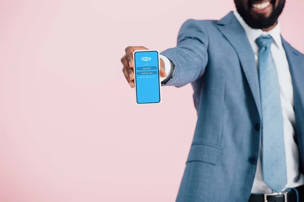 КИЕВ, УКРАИНА - 17 мая 2019 года: обрезанный вид африканского американского бизнесмена в костюме, показывающий смартфон с приложением skype, изолированным на розовом — стоковое фото