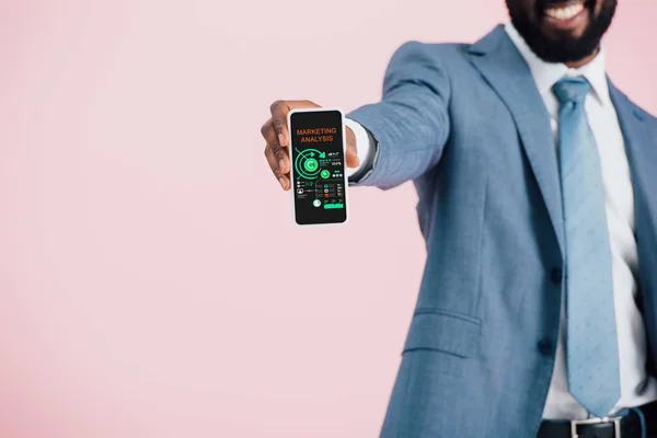 Vista recortada del hombre de negocios afroamericano en traje que muestra el teléfono inteligente con análisis de marketing, aislado en rosa - foto de stock
