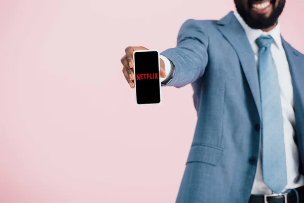 КИЕВ, УКРАИНА - 17 мая 2019 года: обрезанный вид африканского американского бизнесмена в костюме, показывающий смартфон с приложением netflix, изолированным на розовом — стоковое фото
