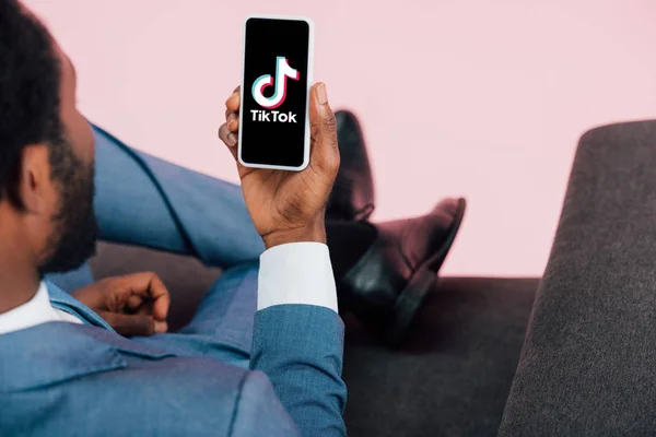 КИЕВ, УКРАИНА - 17 мая 2019 года: обрезанный вид африканского американского бизнесмена, сидящего на кресле и показывающего смартфон с приложением Tik Tok, изолированным на розовом — стоковое фото