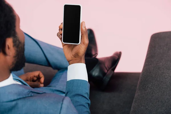 Vista recortada del hombre de negocios afroamericano sentado en sillón y sosteniendo el teléfono inteligente con pantalla en blanco, aislado en rosa - foto de stock