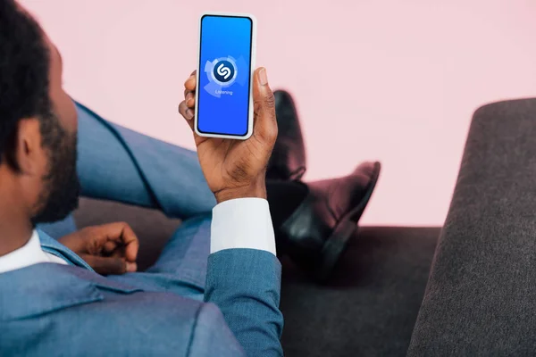 KYIV, UKRAINE - MAY 17, 2019: cropped view of african american businessman sitting on armchair and showing smartphone with shazam app, isolated on pink — Stock Photo