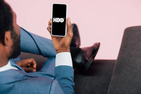 KYIV, UKRAINE - MAIO 17, 2019: visão recortada do empresário afro-americano sentado em poltrona e mostrando smartphone com aplicativo HBO, isolado em rosa — Fotografia de Stock