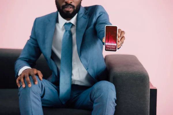 Vista recortada de hombre de negocios afroamericano sentado en el sillón y mostrando el teléfono inteligente con aplicación cursos de comercio, aislado en rosa - foto de stock