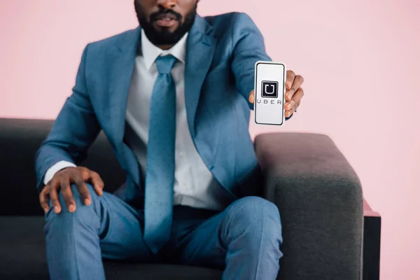 KYIV, UCRAINA - 17 MAGGIO 2019: vista ritagliata dell'uomo d'affari afroamericano seduto sulla poltrona e che mostra smartphone con app uber, isolato su rosa — Foto stock