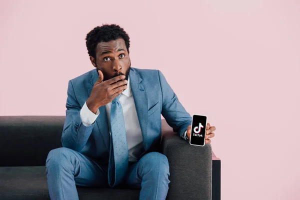 KYIV, UCRANIA - 17 de mayo de 2019: sorprendido hombre de negocios afroamericano en traje sentado en un sillón y mostrando el teléfono inteligente con la aplicación Tik Tok, aislado en rosa - foto de stock