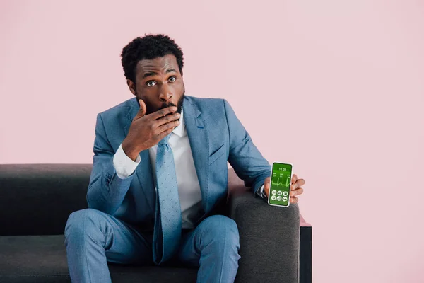 Sorprendido hombre de negocios afroamericano en traje sentado en el sillón y mostrando teléfono inteligente con aplicación de salud, aislado en rosa - foto de stock