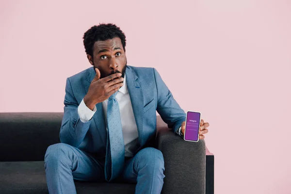 KYIV, UCRANIA - 17 de mayo de 2019: sorprendido hombre de negocios afroamericano en traje sentado en un sillón y mostrando el teléfono inteligente con aplicación instagram, aislado en rosa - foto de stock