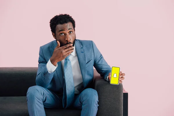 KYIV, UKRAINE - 17 de maio de 2019: empresário afro-americano chocado de terno sentado em poltrona e mostrando smartphone com aplicativo Snapchat, isolado em rosa — Fotografia de Stock