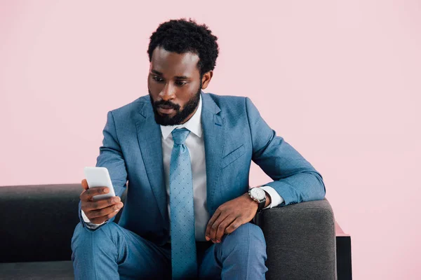 Hombre de negocios afroamericano sentado en un sillón y usando un teléfono inteligente, aislado en rosa - foto de stock