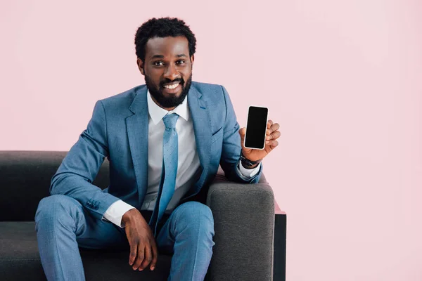 Hombre de negocios afroamericano sentado en sillón y mostrando teléfono inteligente con pantalla en blanco, aislado en rosa - foto de stock