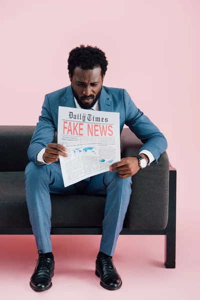 Upset african american businessman reading newspaper with fake news while sitting on sofa, isolated on pink — Stock Photo