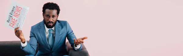Спантеличений афроамериканський бізнесмен тримає газету з фальшивими новинами, сидячи на дивані, ізольовані на рожевому — стокове фото