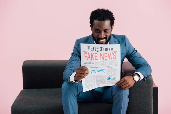 Smiling african american businessman reading newspaper with fake news while sitting on sofa, isolated on pink — Stock Photo