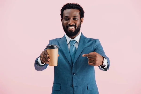 Homme d'affaires afro-américain souriant pointant du doigt le café à emporter, isolé sur rose — Photo de stock