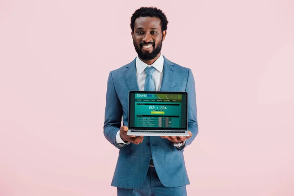 Lächelnder afrikanisch-amerikanischer Geschäftsmann zeigt Laptop mit Sportwetten-Webseite isoliert auf blau — Stockfoto