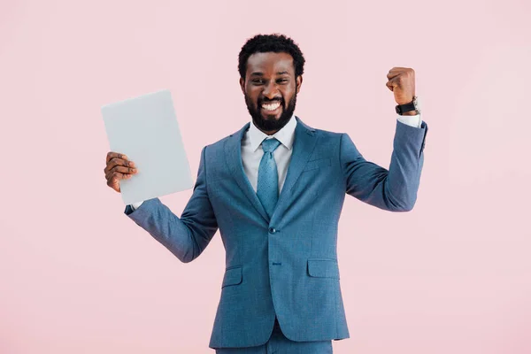 Excited african american businessman in suit holding laptop isolated on pink — Stock Photo