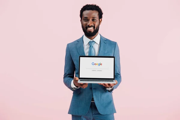 KYIV, UCRANIA - 17 DE MAYO DE 2019: sonriente hombre de negocios afroamericano mostrando portátil con el sitio web de Google, aislado en rosa - foto de stock
