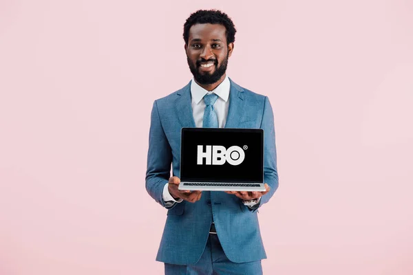 Київ, Україна-17 травня 2019: усміхнений афроамериканець бізнесмен показує ноутбук з сайтом HBO, ізольований на рожевому — стокове фото