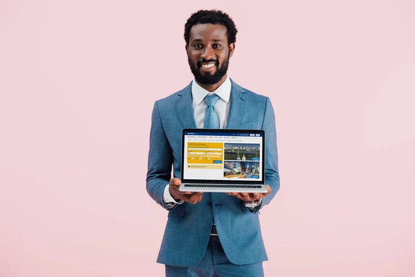 КИЕВ, УКРАИНА - 17 мая 2019 года: улыбающийся африканский бизнесмен, показывающий ноутбук с сайтом бронирования, изолированный на розовом — стоковое фото
