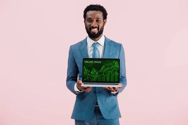 Sonriente hombre de negocios afroamericano mostrando portátil con el comercio en línea aislado en azul - foto de stock