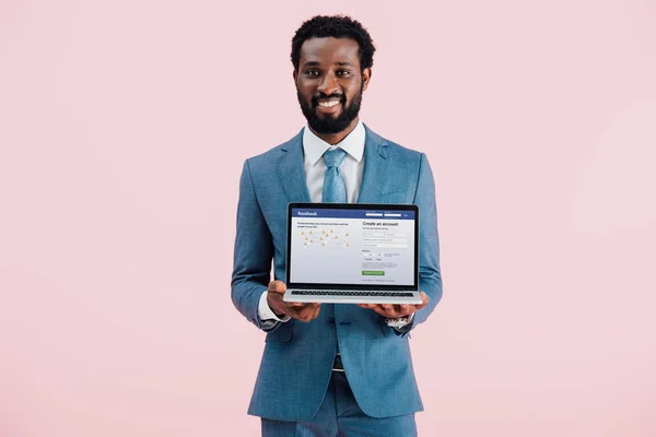 KYIV, UKRAINE - 17 MAI 2019 : homme d'affaires afro-américain souriant montrant ordinateur portable avec site facebook, isolé sur rose — Photo de stock