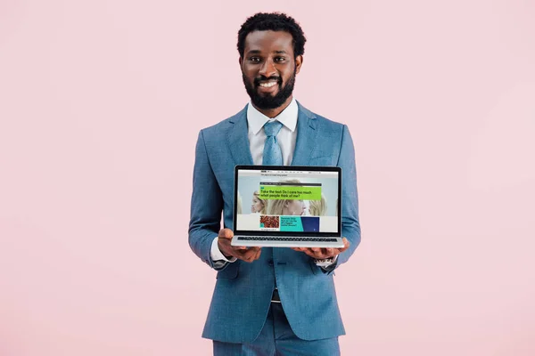 КИЕВ, УКРАИНА - 17 мая 2019 года: улыбающийся африканский бизнесмен показывает ноутбук с сайтом BBC, изолированный на розовом — стоковое фото