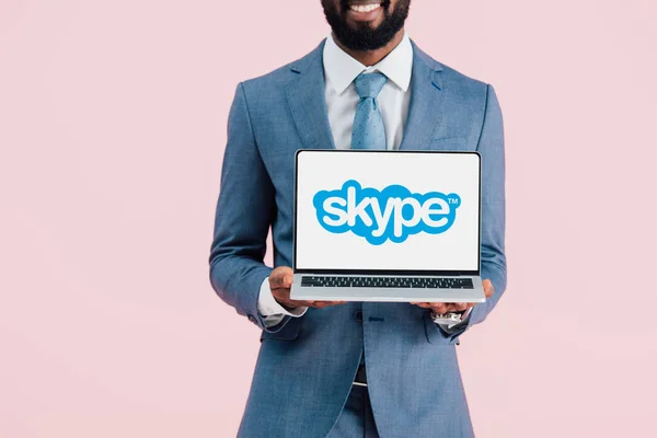 КИЕВ, УКРАИНА - 17 мая 2019 года: обрезанный вид улыбающегося африканского бизнесмена, показывающего ноутбук со скайпом, изолированный на розовом — стоковое фото