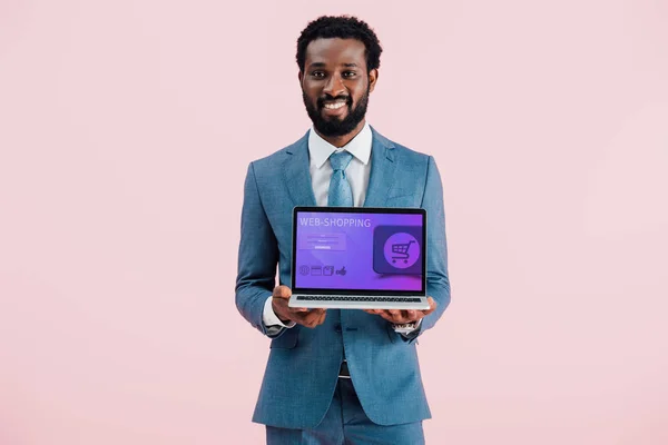 Усміхнений афроамериканський бізнесмен, що показує ноутбук з веб-покупками ізольовано на синьому — стокове фото