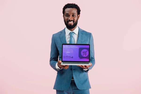 Sonriente hombre de negocios afroamericano mostrando portátil con compras en la web aislado en azul - foto de stock