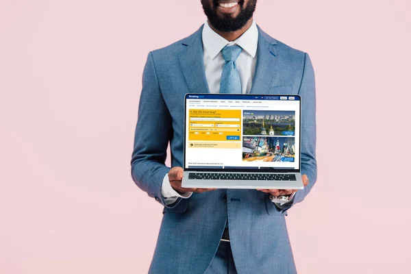 Kyiv, ukraine - 17. Mai 2019: Ausschnittansicht eines lächelnden afrikanisch-amerikanischen Geschäftsmannes, der Laptop mit Buchungswebseite zeigt, isoliert auf rosa — Stockfoto