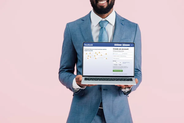 KYIV, UKRAINE - MAY 17, 2019: cropped view of smiling african american businessman showing laptop with facebook website, isolated on pink — Stock Photo