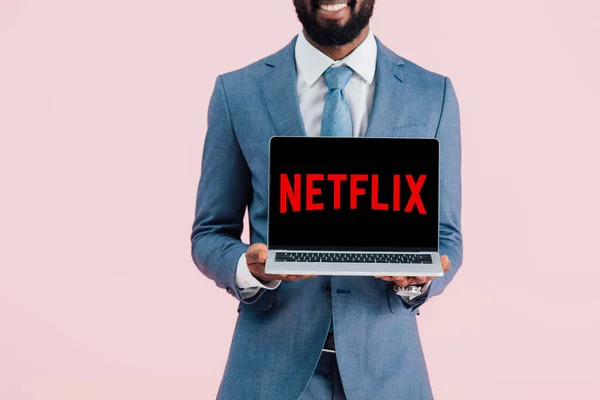KYIV, UCRANIA - 17 de mayo de 2019: vista recortada de un hombre de negocios afroamericano sonriente que muestra el ordenador portátil con el sitio web de netflix, aislado en rosa — Stock Photo