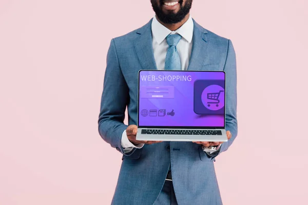 Vista recortada de sonriente hombre de negocios afroamericano mostrando portátil con compras en la web aislado en azul - foto de stock