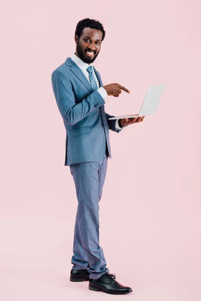 Smiling african american businessman pointing at laptop isolated on pink — Stock Photo