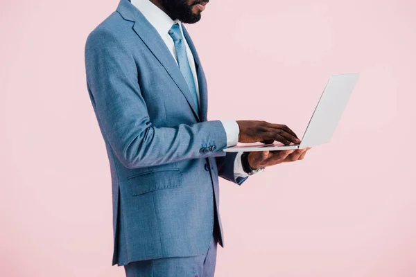Vista recortada de hombre de negocios afroamericano en traje usando portátil aislado en rosa - foto de stock