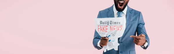 Vista recortada del hombre de negocios afroamericano enojado gritando y leyendo el periódico con noticias falsas aisladas en rosa - foto de stock