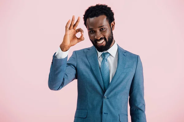 Усміхнений афро-американський бізнесмен у костюмі, що показує знак ОК, ізольований на рожевому — стокове фото