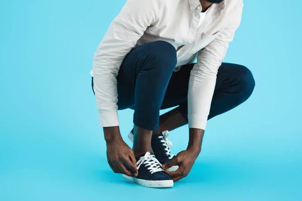 Vista recortada del hombre afroamericano atando cordones en zapatillas aisladas en azul - foto de stock