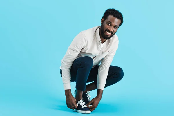 Fröhliche afrikanisch-amerikanische Mann schnürt Schnürsenkel auf Turnschuhen isoliert auf blau — Stockfoto