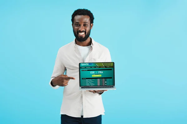 Улыбающийся африканский американец, указывающий на ноутбук со спортивным сайтом, изолированным на синем — стоковое фото