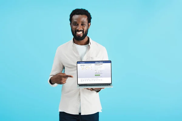 KYIV, UCRANIA - 17 DE MAYO DE 2019: sonriente hombre afroamericano señalando a la computadora portátil con el sitio web de facebook, aislado en azul - foto de stock