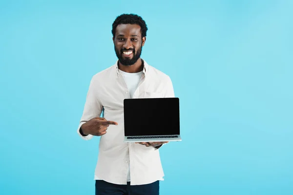 Усміхнений афроамериканський чоловік вказує на ноутбук з порожнім екраном ізольовано на синьому — стокове фото
