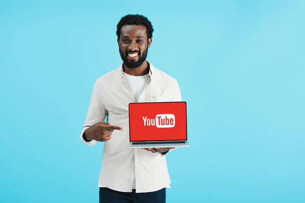 KYIV, UKRAINE - MAIO 17, 2019: homem americano africano sorridente apontando para laptop com site do youtube, isolado em azul — Fotografia de Stock